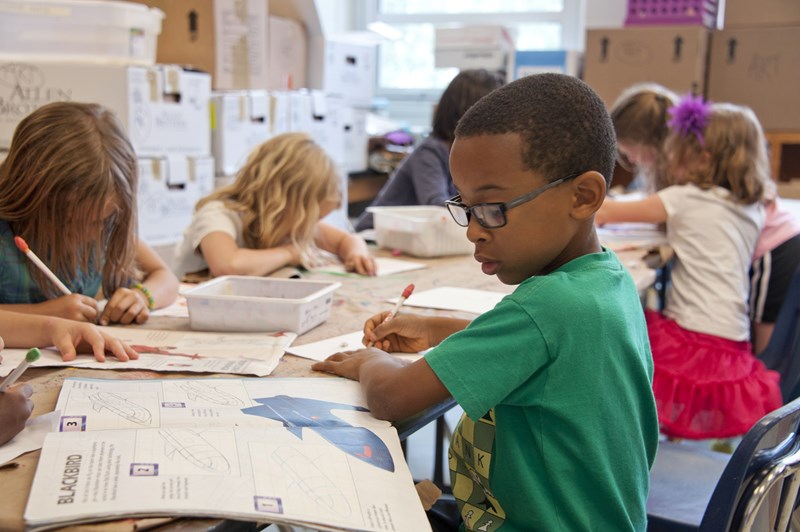 This image concerns equity in education. It showcases a classroom setting with multiple children working from textbooks. The camera’s main focus is on a black child in the foreground who is wearing glasses and a green T-shirt. The child is focusing on his work and has a pen in his hand.
